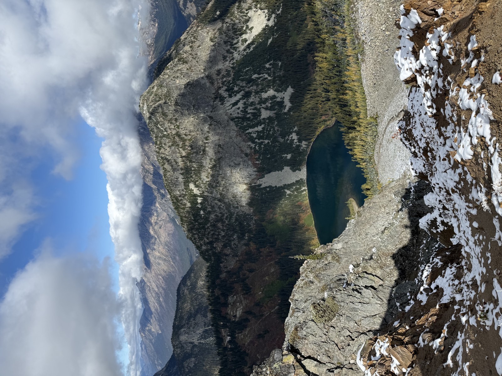 Lake Ann far below a rocky ridge, ringed by golden larches, with snow on the rocks in front