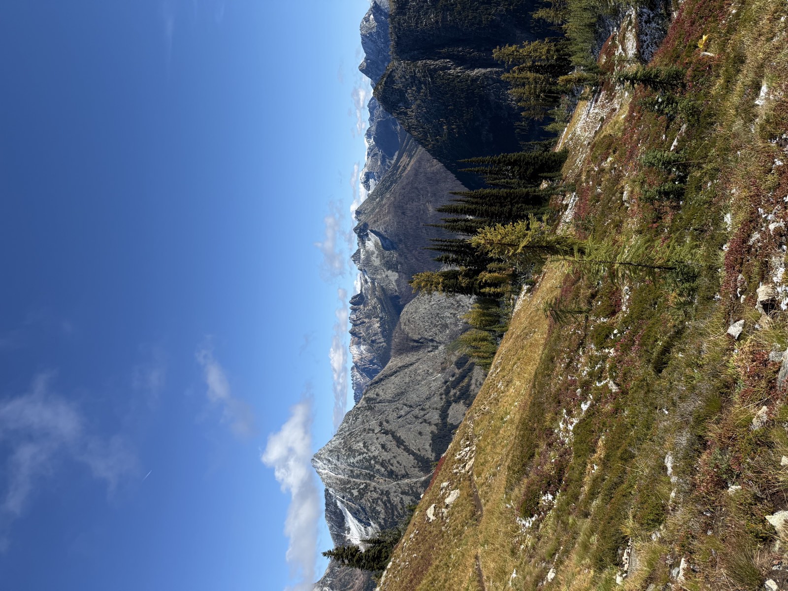 A trail crossing a grassy slope toward a pointed gray peak and distant mountains