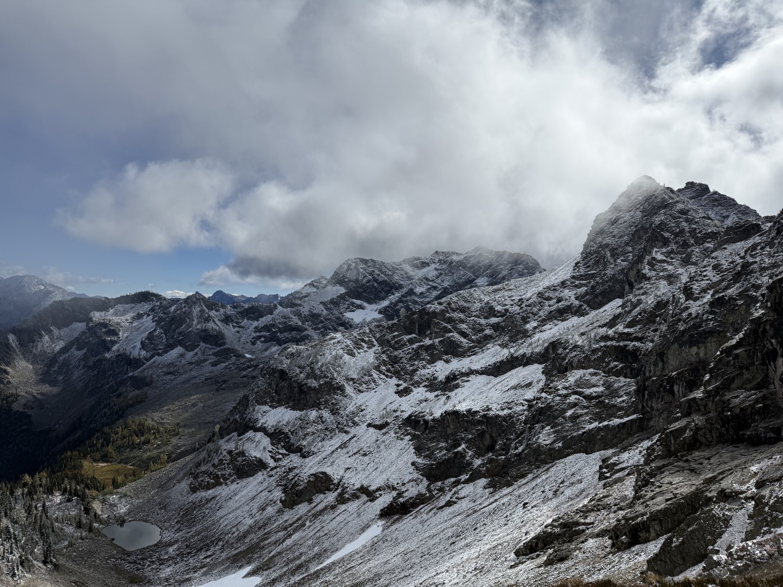 Snow-dusted cliffs and talus above a small tarn, under breaking clouds