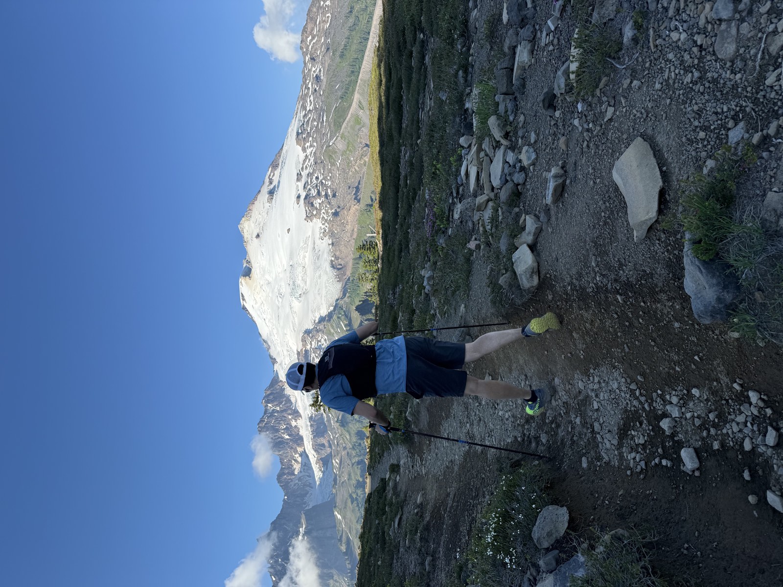 A hiker with trekking poles on a rocky trail through open meadows below Mount Baker