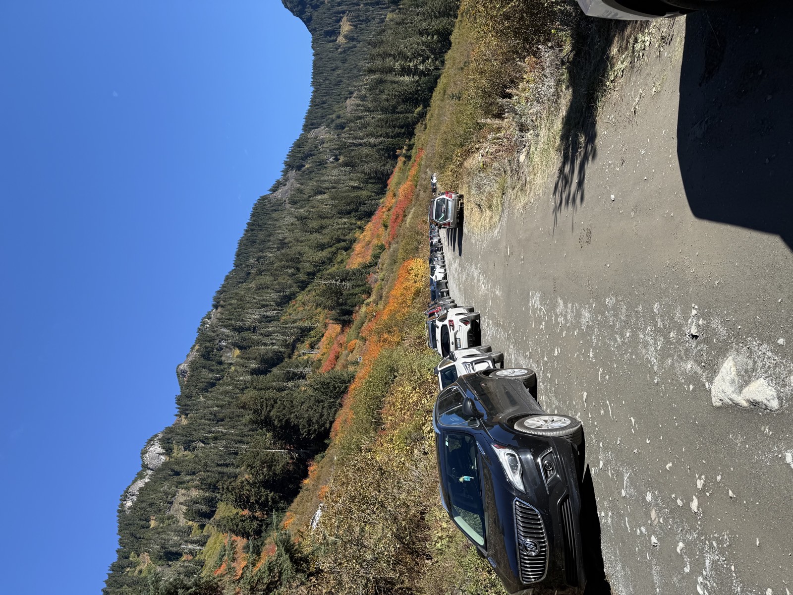 A long line of cars parked along a gravel road below a hillside of orange and red brush